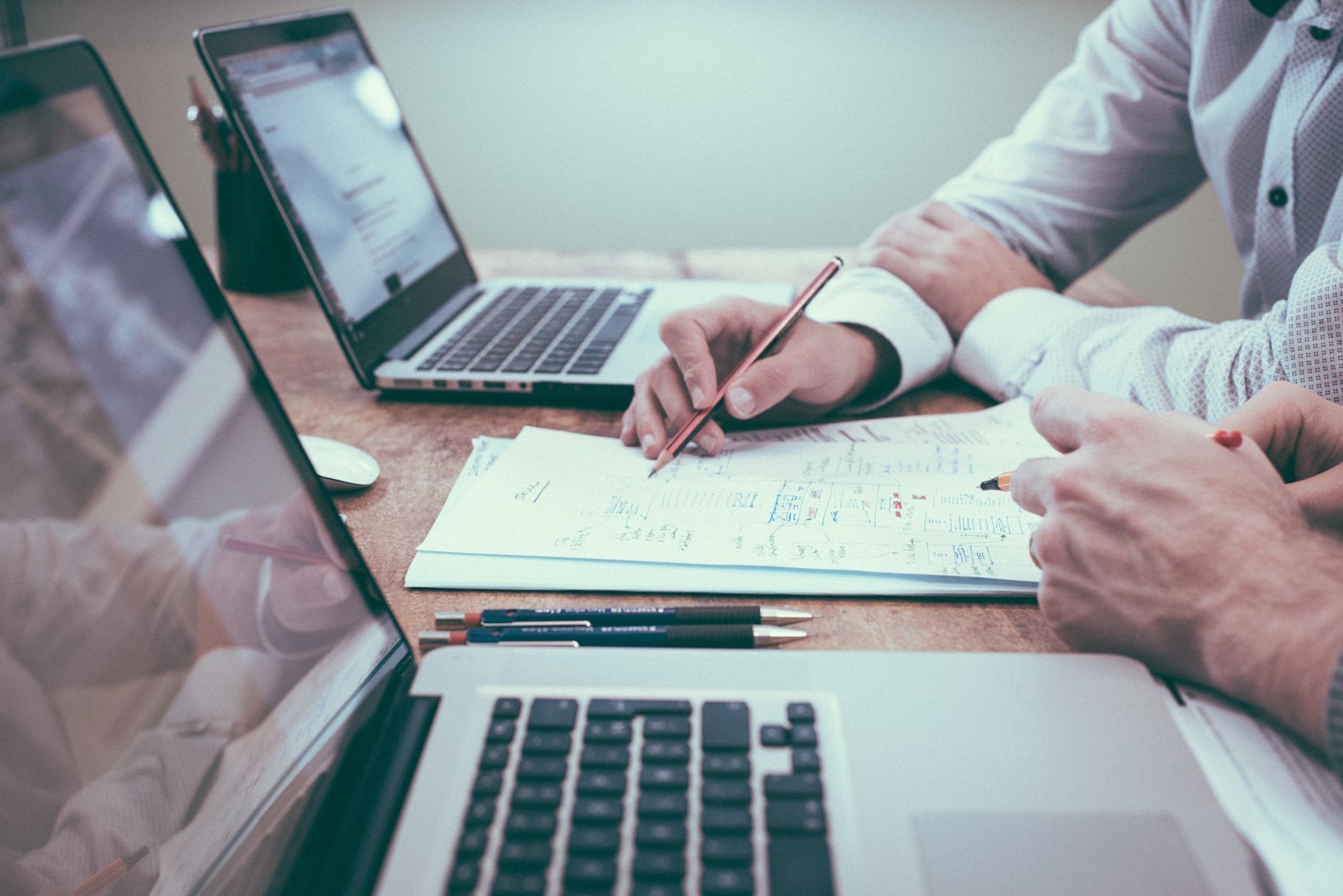 two employees are working together. they both have their laptops open and they have a shared set of notes on the table between them.