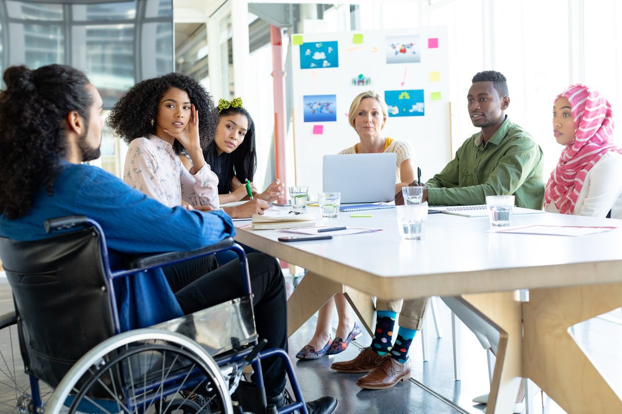 a team meeting between a group of six employees from a range of different ethnicities. The employees are directing their attention to a man in a wheelchair who is seemingly sharing his ideas with the rest of the group. 