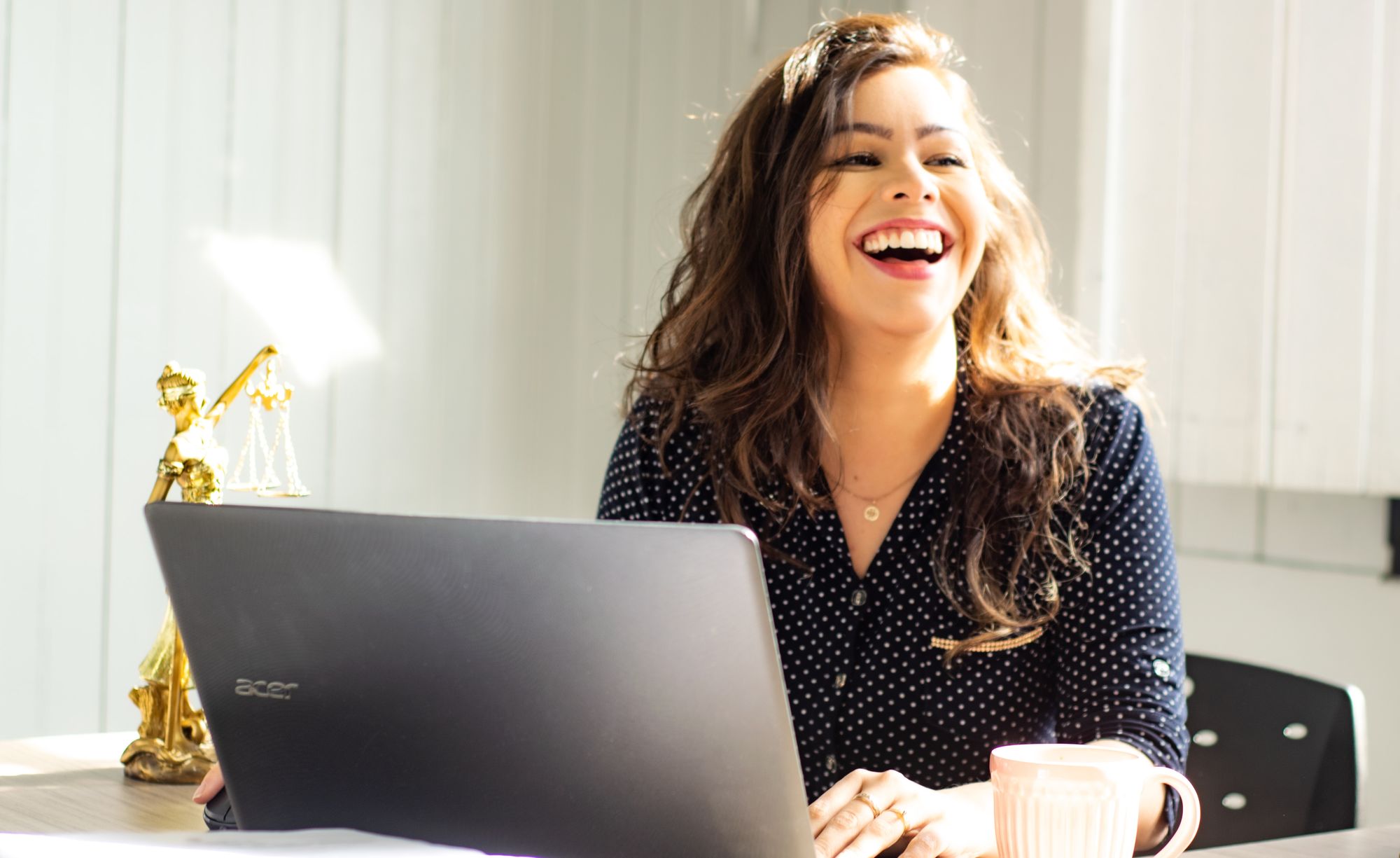 a young brown-haired woman is sat in front of her laptop, looking away and laughing