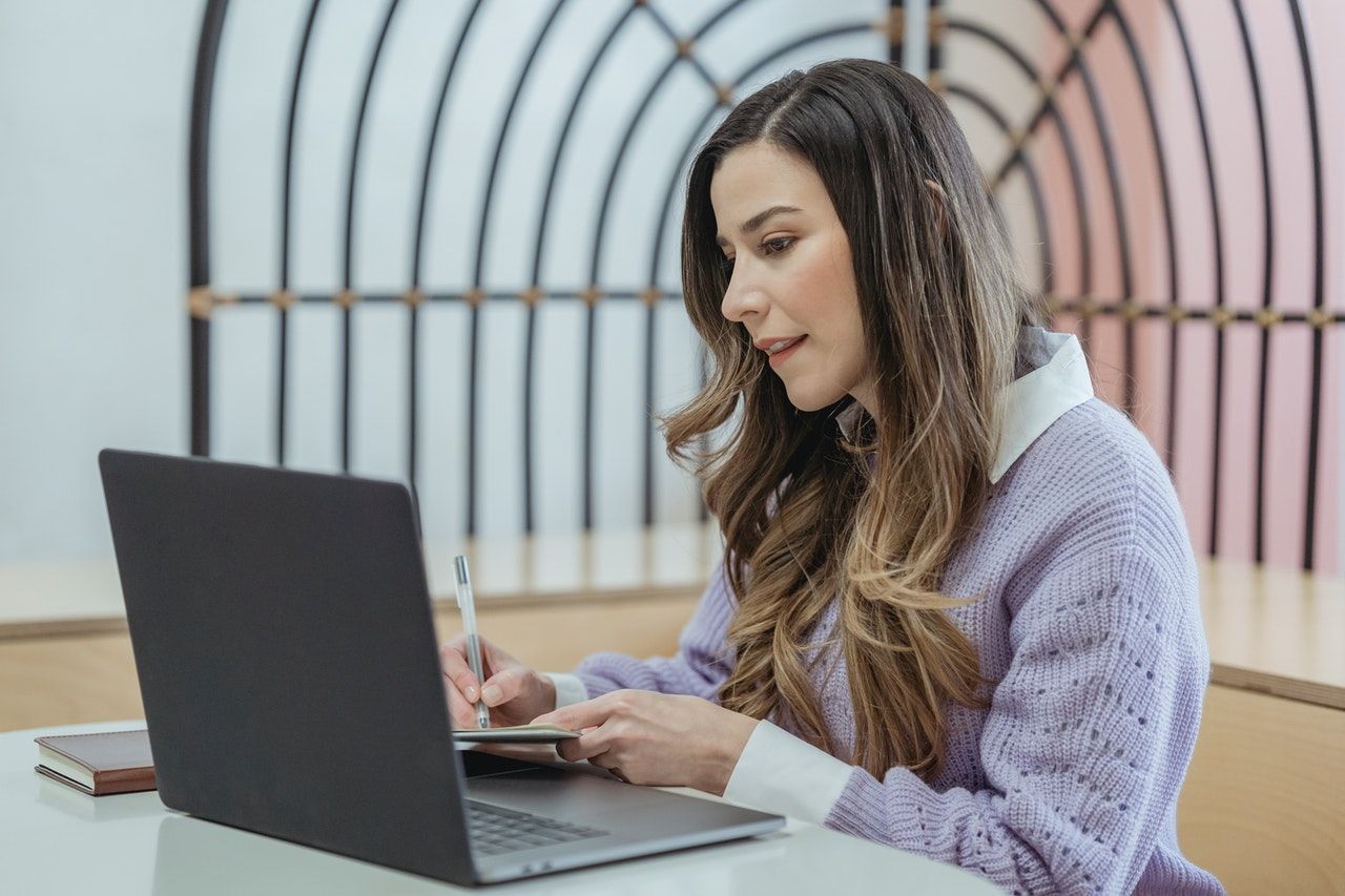 A young woman is focusing on her laptop whilst also taking notes.