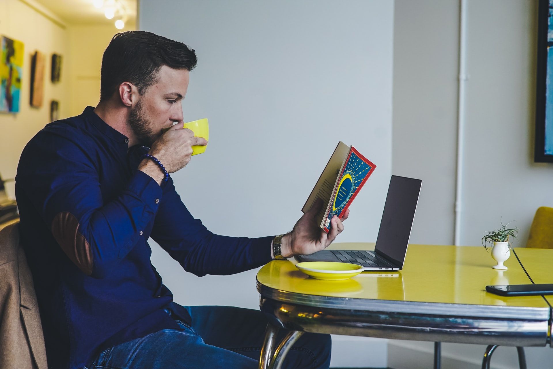 A remote worker sits at his desk, drinking a coffee and reading a book. His laptop is open in front of him.