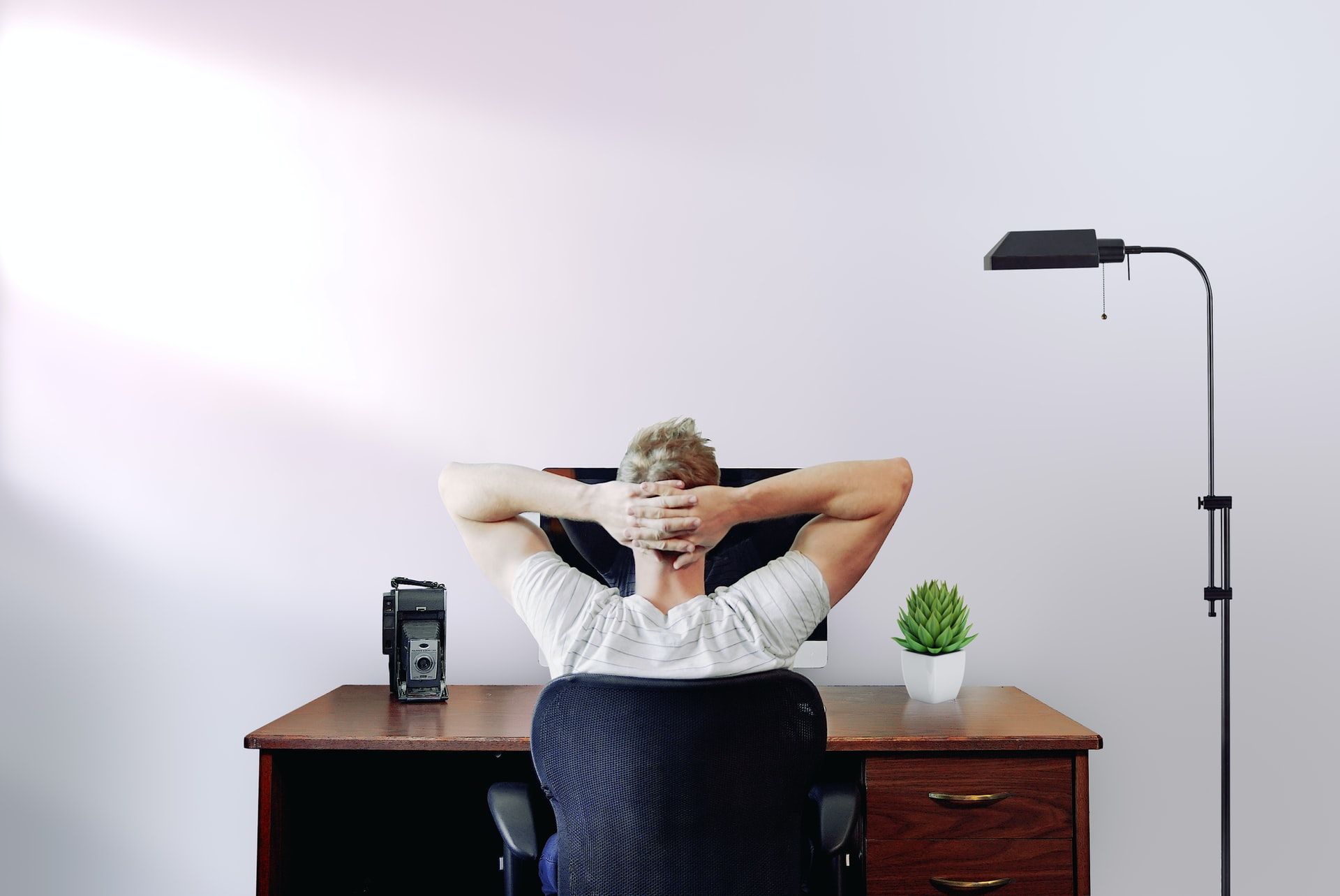 A remote employee sits at his home workspace with his hands behind his head and his desktop computer in front of him.