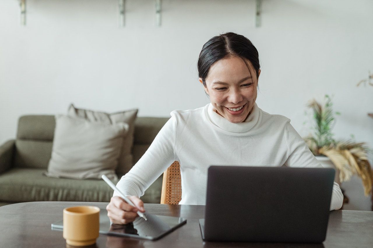 A remote worker is sat at her desk with her laptop open and taking notes. She's smiling at her screen as though she's on a video call.